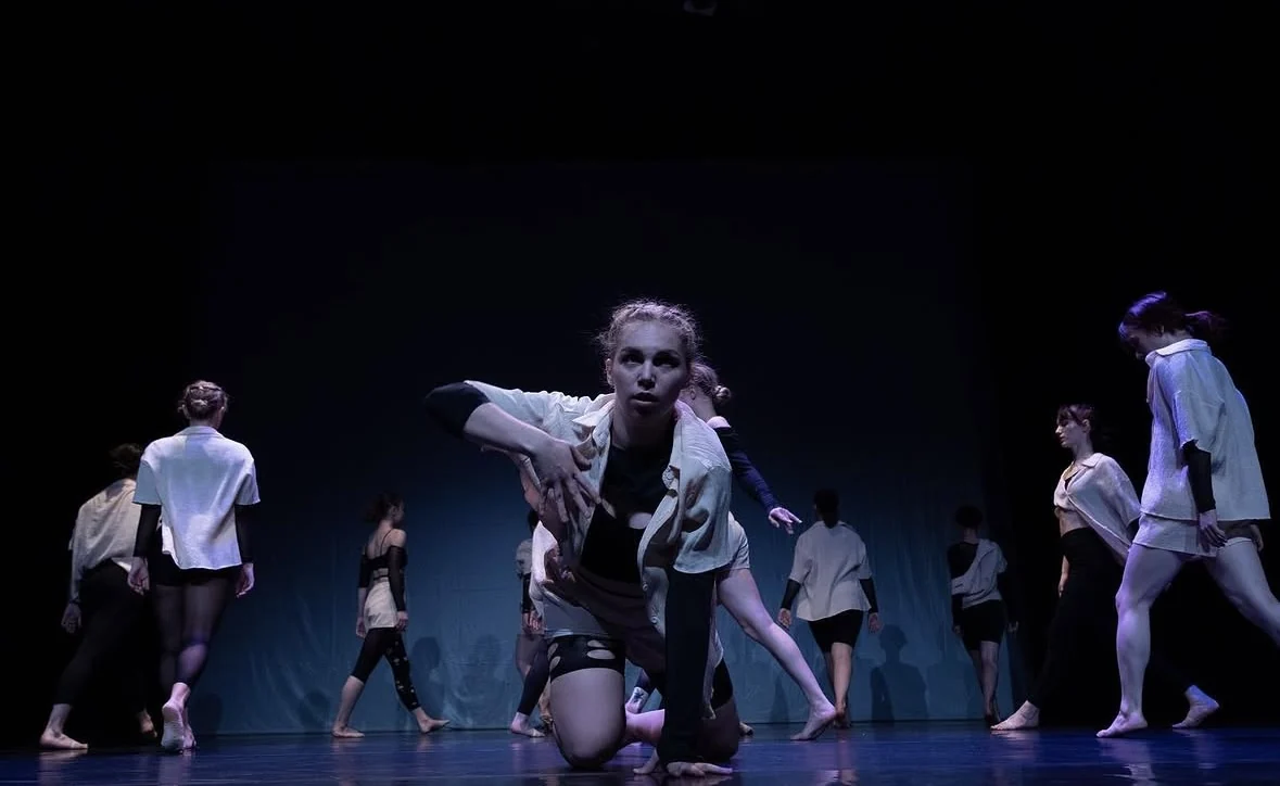 An ensemble performing on a dark stage, with a dancer reaching forward at the centre
